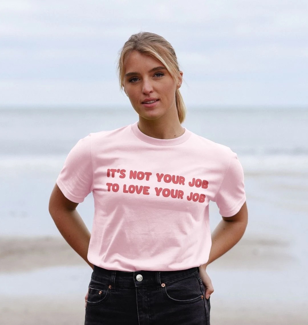 Model wearing a light pink t-shirt with a large dark pink text along the front on two lines reading It's Not Your Job To Love Your Job.